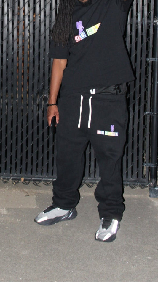 Person wearing a black outfit with a logo in front of a black metal gate at night.